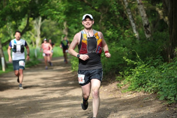 Runner in the foreground smiling at the camera under tree canopy on a wide trail. Other runners are out of focus in the background heading the same way